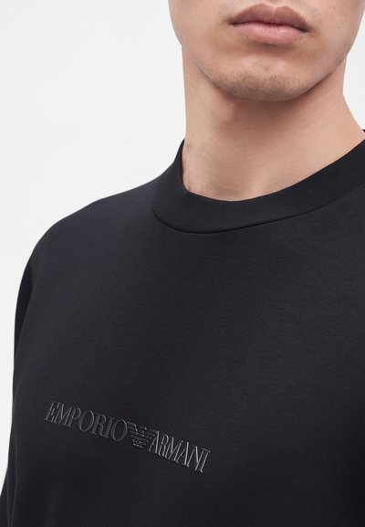 Gros plan d'une personne portant un t-shirt noir avec le texte en relief "EMPORIO ARMANI" et le logo sur la poitrine.