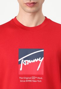 Červené bavlnené tričko s grafickým logom v modrej, bielej a červenej farbe. Zaujímavé detaily zahŕňajú okrúhly výstrih a hladkú textúru.