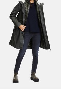 Tmavě zelený nepromokavý kabát s prošívanou vnitřní podšívkou, šedou kapucí, zapínáním na zip a dvěma předními kapsami, nošený přes námořnicky modrý svetr a džíny.