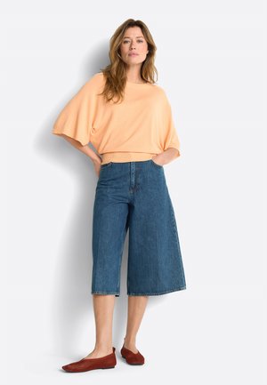 Kobieta stoi z rękami w kieszeniach, ubrana w brzoskwiniowy top z szerokimi rękawami, niebieskie dżinsowe culottes i czerwone plecione baleriny na jasnym tle.