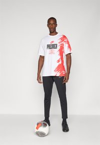 Hvid t-shirt med rød grafisk design, "PREMIER FOOTBALL," sammen med sorte bukser og sorte sko, med en fodbold i nærheden.