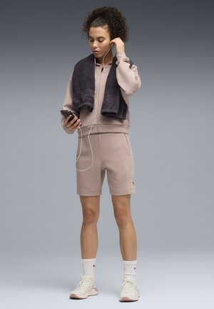 Jeune femme en short de sport beige, sweat-shirt, baskets, chaussettes blanches et une serviette foncée écoutant de la musique avec des écouteurs reliés à un smartphone.