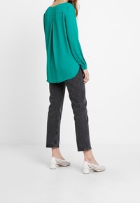 Blusa lunga teal con orlo arrotondato, abbinata a jeans neri skinny e scarpe bianche con tacco chunky caratterizzate da ritagli.