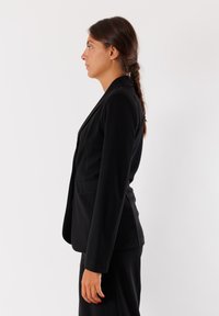 Blazer nero realizzato in tessuto liscio, con vestibilità su misura, revers a becco di flauto e una tasca frontale singola, mostrato di lato.