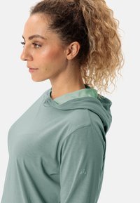 Helle grüne Kapuzenjacke mit weicher Textur, ausgestattet mit einer Kapuze und einer kontrastierenden inneren Fütterung. Einfaches Design mit einem Logo auf dem Ärmel.