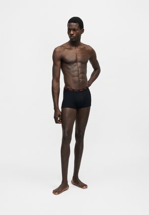Homme torse nu, debout pieds nus, portant des boxer noirs avec le logo rouge "HUGO" sur la ceinture, sur fond blanc uni.