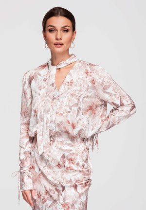 Femme portant un chemisier à fleurs pastel avec un nœud au cou et une jupe assortie, posant d'une main sur la hanche devant un fond uni.