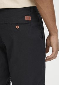 Zwarte shorts van een gestructureerde stof, met een bruine leren merkpatch en een knoopdetail op de achterzak. Subtiele stiksels aanwezig.