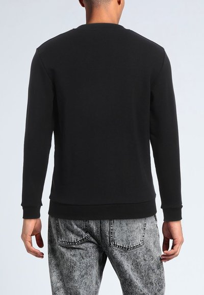 Felpa nera con maniche lunghe, collo rotondo, polsini e orlo a coste, realizzata in misto cotone, indossata con jeans grigi chiari effetto acido.