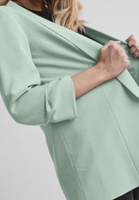 Blazer vert menthe à coupe décontractée, avec manches 3/4 et ouverture avant. Tissu lisse avec une texture subtile, sans fermeture.