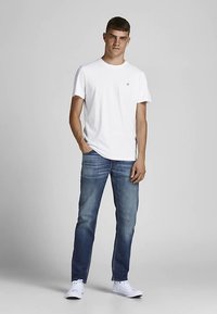 Biały t-shirt z krótkim rękawem z małym logo, zestawiony z ciemnoniebieskimi dżinsami i białymi trampkami, stojący na neutralnym tle.