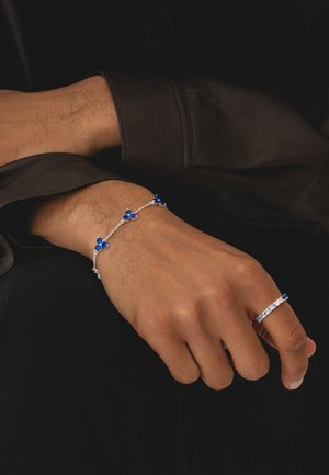 Mano con anello d'argento con pietre blu e delicato braccialetto d'argento abbinato con ciondoli a forma di fiore blu su sfondo di tessuto scuro.