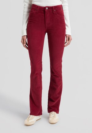 Femme portant un pantalon en velours côtelé rouge foncé taille haute et des baskets blanches, debout devant un fond clair uni.