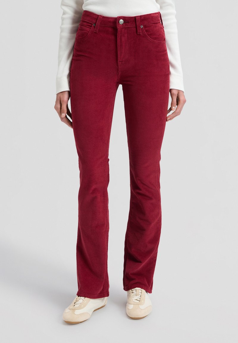 Femme portant un pantalon en velours côtelé rouge foncé taille haute et des baskets blanches, debout devant un fond clair uni.