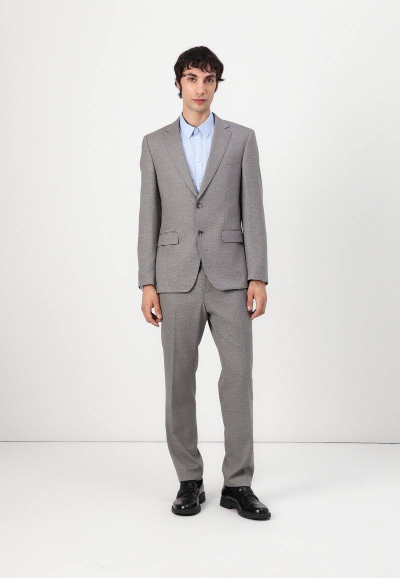 Completo grigio su misura con giacca a un bottone, pantaloni dritti e camicia azzurra chiara. Scarpe nere completano l'outfit.