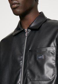 Zwarte leren jas met kraag, ritssluiting en een borstzak met een logopatch. Gladde textuur en een aansluitend ontwerp.