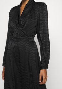 Mustapukuinen satiinimekko, jossa on strukturoidusta viuhkakuosisesta kangasta, pitkät hihat, V-mallinen kaula-aukko ja kaksi nappikoristetta vyötäröllä.