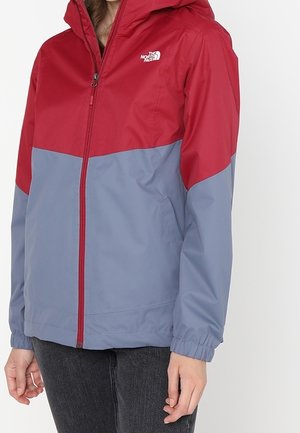 Donna con giacca con cappuccio North Face rossa e grigia chiusa fino in alto, abbinata a pantaloni grigio scuro, in piedi davanti a uno sfondo bianco.