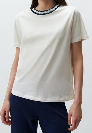 Femme portant un t-shirt blanc à manches courtes avec un col festonné noir et blanc et un pantalon bleu foncé.