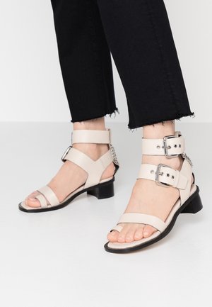 Nohy obuté v béžových sandálech s otevřenou špičkou a dvěma sponkovými řemínky na kotník, doplněné o kalhoty s roztrhaným lemem v černé barvě.