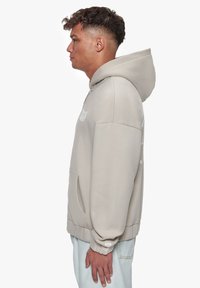 Beige hoodie med en frontlomme og snøre i hætten. Har et logo på brystet, ribbede mansjetter og kant, lavet af blødt stof.