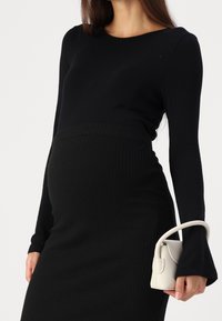 Černé žebrované šaty s dlouhými rukávy, přiléhavý střih zvýrazňující těhotenskou siluetu; drží malou světle zbarvenou kabelku.