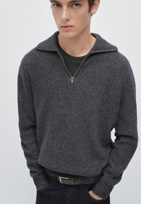 Pull gris en maille côtelée avec un col demi-zip, tissu texturé, manches longues et ourlet ajusté. Porté par-dessus un t-shirt noir.