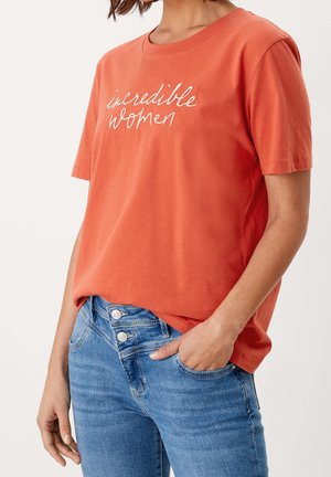 Rustfarvet bomulds T-shirt med korte ærmer, der har broderet teksten "incredible women". Sammen med lyseblå jeans.