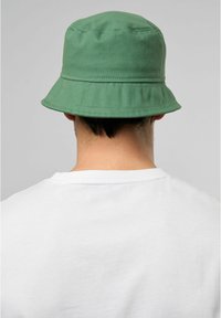 Grön bomulls-bucket hat med bred skärm, slät yta och sydda detaljer, visad bakifrån ovanför en vit t-shirt.