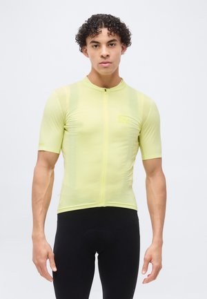 Νεαρός με σγουρά μαλλιά, φοράει εφαρμοστό κίτρινο ποδηλατικό φανελάκι με κοντά μανίκια και μαύρο ποδηλατικό σορτς, στέκεται μπροστά σε απλό φόντο.