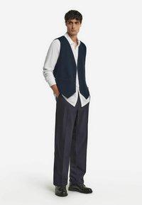 Gilet blu navy con chiusura a bottoni, indossato su una camicia bianca; abbinato a pantaloni scuri e suole nere. Tessuto dalla consistenza liscia.