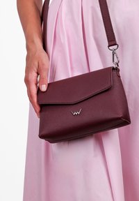 Burgundská texturovaná kabelka přes rameno s klopou a stříbrným logem. Příložený ramenní popruh s kovovou sponou pro snadné nošení.