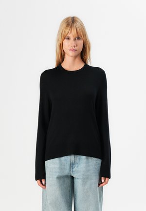 Donna con capelli biondi lisci che indossa un pullover nero a maniche lunghe e jeans a gamba larga di colore blu chiaro, in piedi contro uno sfondo bianco.