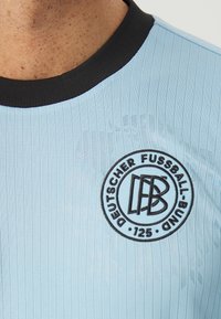 Ljusblå sporttröja med ribbad textur, svart krage och cirkulär svart emblem med "Deutscher Fussball-Bund" och "125."