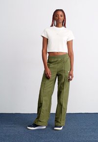 Hvit cropped t-skjorte, olivengrønne vide bukser med sømdetaljer og mørke sneakers. Stoffet ser glatt ut med en atletisk passform.