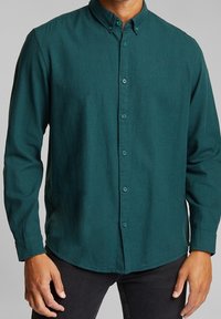 Camisa verde oscuro de manga larga con botones, confeccionada en tejido texturizado, con dobladillo redondeado y cuello abotonado. Botones a juego.