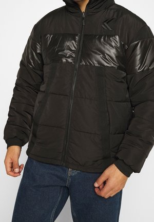 Man draagt een zwarte gewatteerde jas met doorgestikte en glanzende panelen, een ritssluiting aan de voorkant en elastische manchetten, gecombineerd met blauwe spijkerbroek.