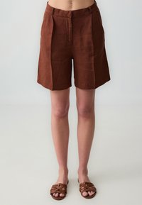 Pantaloni corti in lino marrone con una vestibilità sartoriale, dotati di cintura e pieghe frontali. Abbinati a sandali intrecciati marroni.