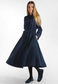Abito camicia blu navy a maniche lunghe con vita stretta, gonna ampia e una leggera texture, abbinato a ballerine scure e collant neri.