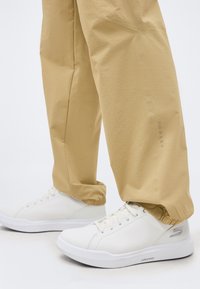 Άτομο που φοράει μπεζ παντελόνι Oakley και λευκά αθλητικά παπούτσια Skechers Arch Fit σε καθαρό λευκό δάπεδο.