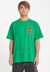 T-shirt en coton vert à manches courtes et col rond. Présente trois motifs imprimés en orange, noir et jaune sur le devant.