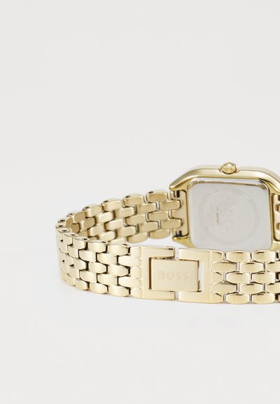 Montre-bracelet dorée avec un boîtier rectangulaire, un bracelet en maille et un fermoir avec le logo "BOSS" gravé. Surface métallique lisse, design minimaliste.