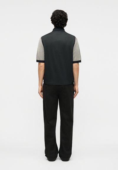 Άνδρας με σκουρόχρωμα, κατσαρά μαλλιά που φοράει μαύρο γιλέκο πάνω από ριγωτό κοντομάνικο πουκάμισο και μαύρο παντελόνι, στέκεται γυρισμένος με την πλάτη σε απλό φόντο.