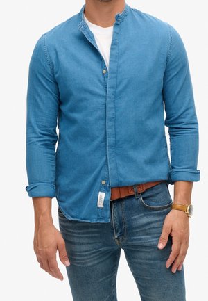 Hombre con una camisa azul abotonada sobre una camiseta blanca, vaqueros azules con un cinturón marrón y un reloj de pulsera dorado en la muñeca izquierda.