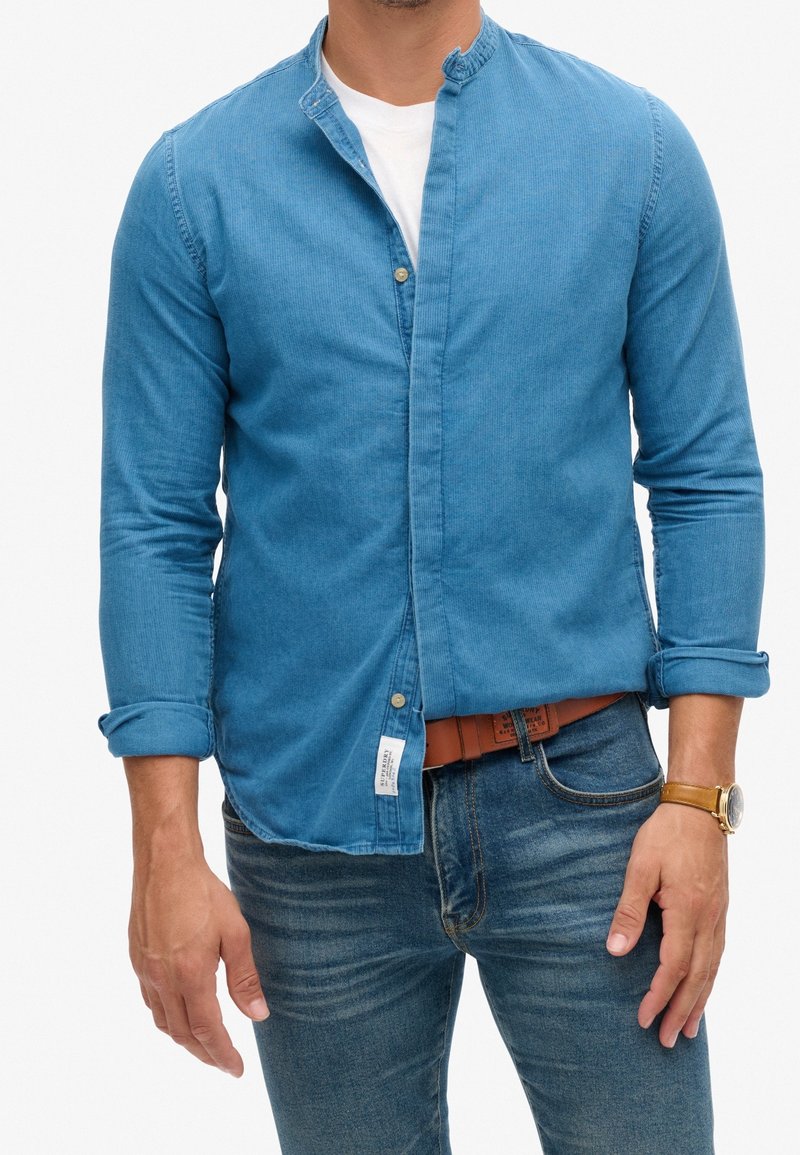 Homme portant une chemise bleue à boutons sur un t-shirt blanc, un jean bleu avec une ceinture marron et une montre-bracelet en or au poignet gauche.
