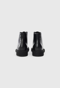 Botas de tobillo de cuero negro con una textura suave, puntera redondeada y suelas de goma negras en contraste. Detalles de costura en la parte trasera.