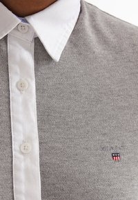 Polo de punto gris con cuello blanco, presenta pequeños botones y un logo bordado en el pecho. Textura suave y diseño casual.