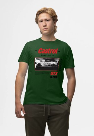 Giovane uomo con capelli biondi che indossa una t-shirt grafica da corsa Castrol GTX verde scuro e pantaloni scuri, in piedi con una mano in tasca.