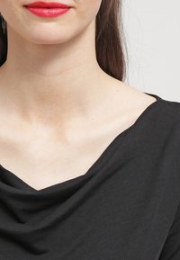 Top nero drappeggiato con scollatura ampia, texture liscia e tessuto morbido. Presenta un movimento sottile lungo le spalle e l'area del colletto.