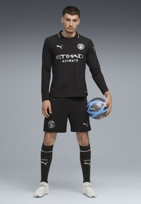 Mandlig atlet står med en blå og sort fodbold i hånden, iført sort Manchester City fodboldtøj, hvide støvler og sorte sokker.